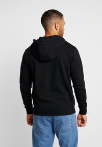 Sort hættejakke med snoretræk i hætten, lange ærmer og ribbede manchetter, parret med lyseblå denimjeans. Stoffet ser blødt og struktureret ud.