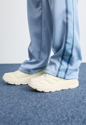 Zapatillas deportivas blancas con malla texturizada y superposiciones lisas, combinadas con pantalones azul claro que tienen dos franjas verticales más oscuras.