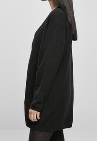 Cardigan long noir en tricot texturé, coupe ample, épaules tombantes et poches latérales. Présente un col rond et des poignets côtelés.