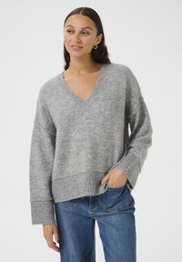 Pulover tricotat gri cu decolteu în V, umeri lăsați și manșete cu nervuri. Textură moale; marginea este ușor scurtată. Asortat cu blugi denim albastri.