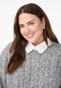Grå kabelstrikket sweater med rund hals, båret over en hvid skjorte med dekorative detaljer; glat hår og neutral baggrund.