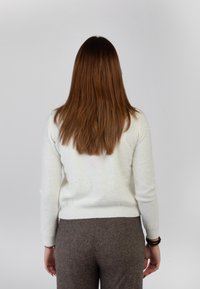 Maglione chiaro in crema con piccole decorazioni glitterate, polsini e orlo a costine. Abbinato a pantaloni marroni con texture. Vista posteriore con capelli lunghi e marroni.