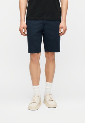 Mężczyzna ubrany w ciemnoniebieskie krótkie spodenki do kolan, białe skarpetki, kremowe trampki i czarną koszulkę, stojący na jednolitym tle.