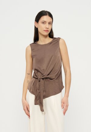 Femme aux longs cheveux foncés portant un haut marron sans manches noué à la taille et un pantalon blanc, debout devant un fond clair uni.