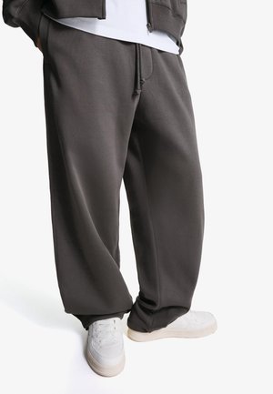 Person trägt lockere dunkelgraue Jogginghose mit Kordelzug und weiße Sneakers, steht vor einfachem hellem Hintergrund.