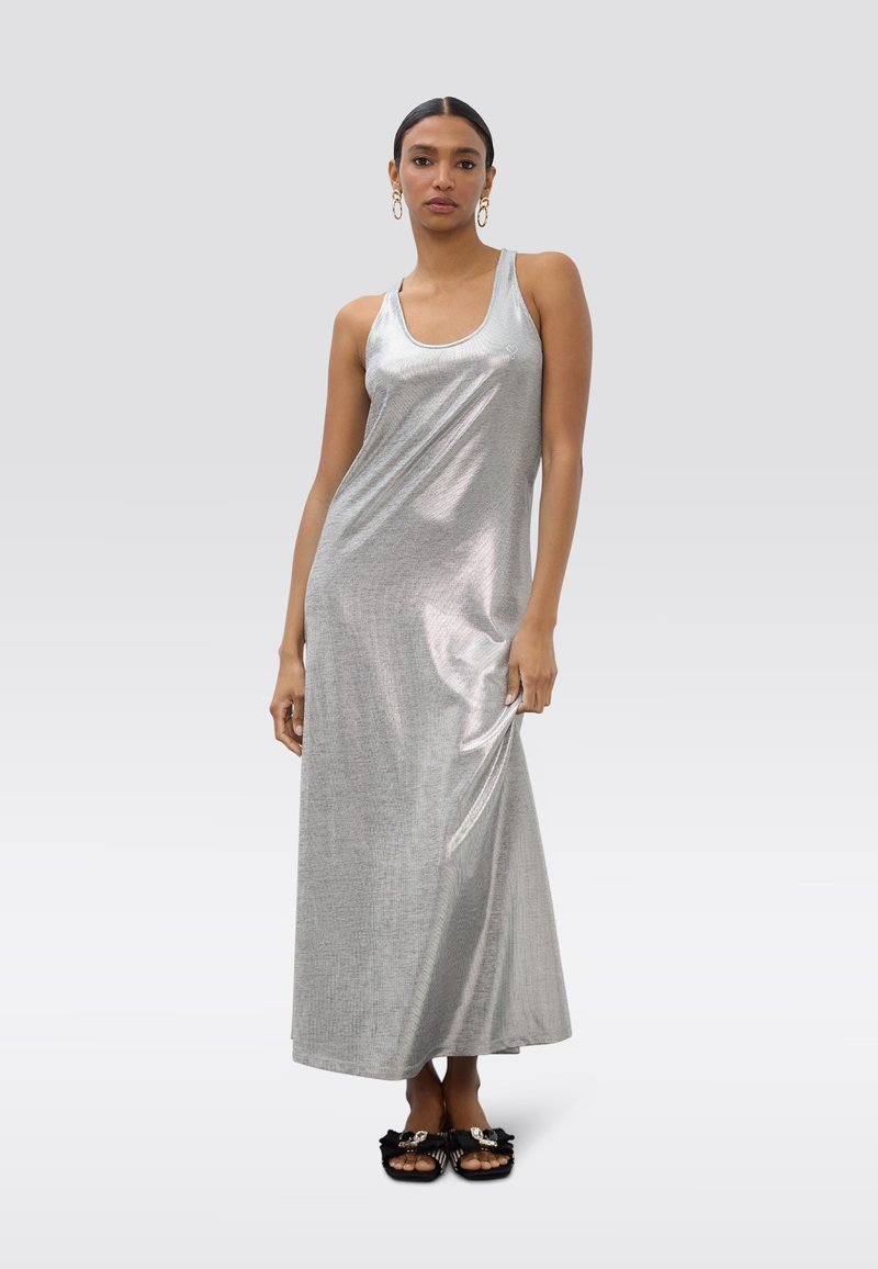 Robe longue argentée avec un col rond et des bretelles fines, présentant une texture lisse et brillante. Associée à des sandales noires ornées de nœuds.