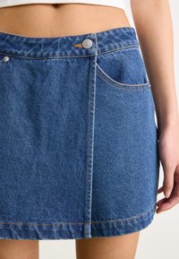Μπλε τζιν μίνι φούστα με μπροστινό κλείσιμο κουμπιού, πλάγιες τσέπες και αντίθεση ραφής κατά μήκος του στριφώματος και των άκρων των τσεπών.