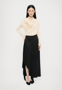 Beige draperige top met lange mouwen, gecombineerd met een zwarte asymmetrische maxirok. De rok heeft een vloeiende silhouet. Zwarte spitse hakken maken de look compleet.