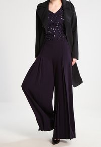 Svart trenchcoat över en mörkt lila topp med ett mönstrat design, kompletterad med vida lila byxor med plisseringar.