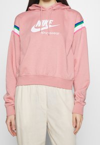 Růžová mikina Nike sportovního stylu, která má zkrácený střih, bílé logo a barevné pruhy na rukávech. Měkká tkanina s volným střihem.