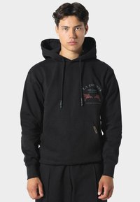 Joven vestido con una sudadera negra con capucha, que tiene un bolsillo frontal y un pequeño texto gráfico e ilustraciones en el pecho izquierdo.