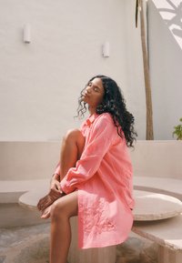 Ilga rožinė marškinėliai su siuvinėtais gėlių raštais, lengva medžiaga, laisvas kirpimas, sagomis užsegami priekyje. Modelis sėdi ant akmeninio stalo.