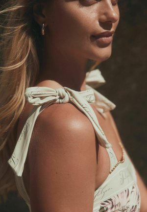 Mladá žena s viazanými kvetinovými ramienkami šiat cez plecia, nosiaca zlaté kruhové náušnice, osvetlená teplým slnečným svetlom.