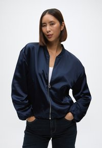 Jūras zila satīna bomber jaka ar ribotu apkakli, rāvējslēdzēju, sānu kabatām un elastīgām piedurknēm, savienota ar tumšiem džinsiem.
