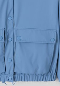 Veste imperméable bleu clair avec une texture lisse, boutons-pression, deux poches avant et un ourlet élastiqué pour un ajustement sécurisé.