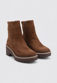 Botas de tobillo de ante marrón con un tacón grueso y suela acanalada. Presenta una cremallera lateral y costuras en contraste a lo largo de las costuras.