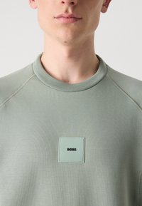 Světl zelená mikina s vroubkovaným kulatým výstřihem, s čtvercovou visačkou s nápisem "BOSS" v černé. Hladký povrch a jednoduchý design.