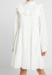 Vit klänning med volangkrage och ärmar, texturerat tyg, långa ärmar, knappstängning och flerlagd fåll. Har en löst sittande design.