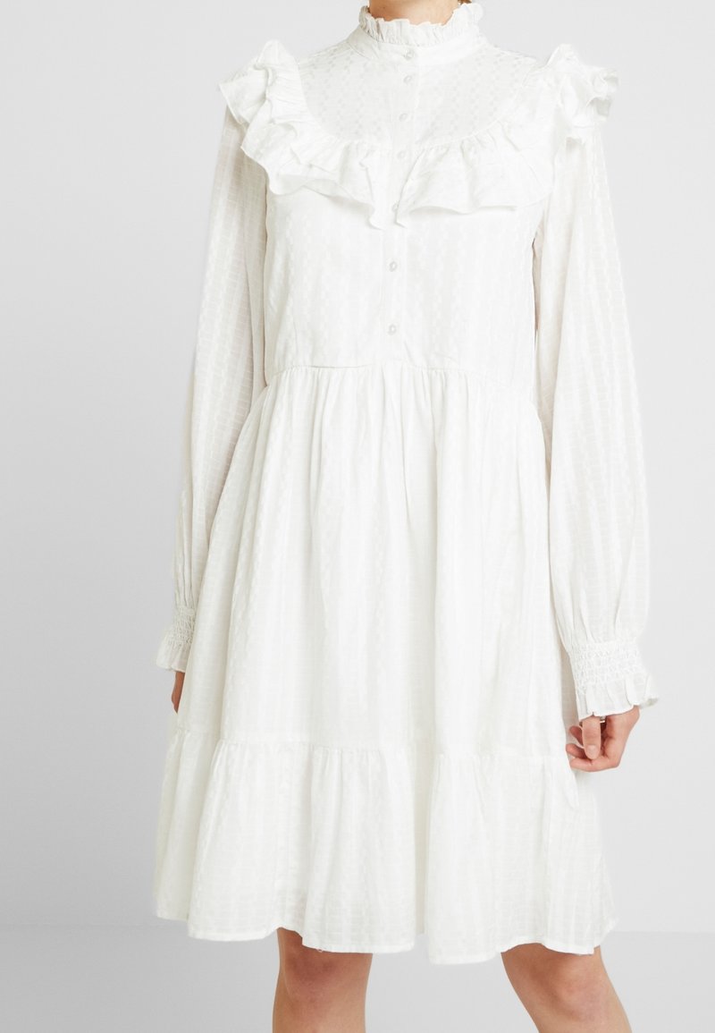 Vit klänning med volangkrage och ärmar, texturerat tyg, långa ärmar, knappstängning och flerlagd fåll. Har en löst sittande design.