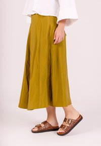Una lunga gonna fluida in tessuto verde oliva con una sottile trama, abbinata a sandali marroni con motivi a intaglio e dettagli con fibbie.