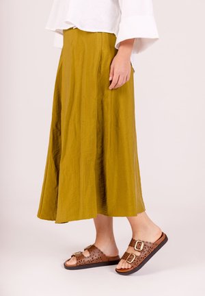 Une longue jupe fluide en tissu vert olive avec une texture subtile, associée à des sandales marron ornées de découpes et de boucles.