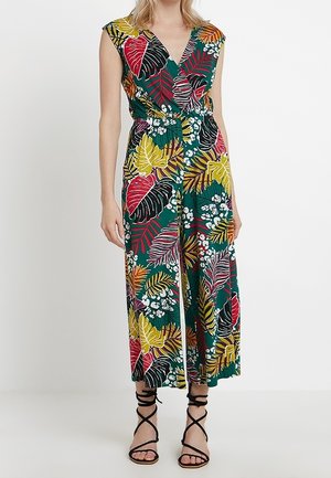 Combinaison sans manches à col en V avec un imprimé multicolore de feuilles tropicales, jambes larges, et sandales noires à lanières, portée par une personne debout.