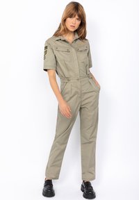 Mono utilitario beige con mangas cortas, bolsillos en el pecho y detalle de cinturón. Presenta una insignia en el brazo izquierdo y piernas ajustadas en forma de cono.