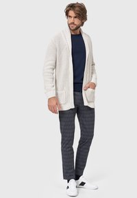 Cardigan lavorato a maglia color crema con colletto a scialle, dotato di due tasche frontali, indossato sopra un maglione blu navy e pantaloni a quadri. Sneakers bianche completano il look.
