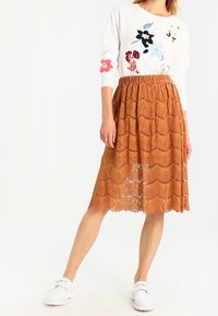 Lace midi-nederdel i rustfarve med klokkeformet kant, parret med en hvid sweater med blomsterbroderi. Hvide sneakers fuldender looket.