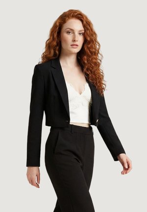 Donna con lunghi capelli rossi ricci indossa un blazer nero corto sopra un top con pizzo bianco e pantaloni neri a vita alta, in piedi su sfondo bianco.