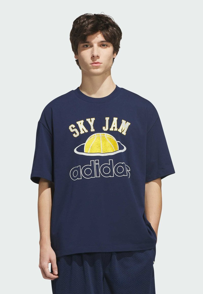Jeune homme aux cheveux bouclés foncés portant un t-shirt Adidas bleu marine "Sky Jam" avec un graphisme jaune de basketball.