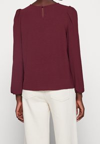 Blouse bordeaux à manches longues, en tissu texturé, avec un détail dos en forme de goutte, associée à un pantalon large de couleur blanc cassé.