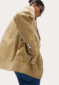 Svetle hnědá semišová bunda s boxy střihem, featuring stříbrné zipy na rukávech a knoflíkovou aplikaci na spodním lemu. Texturovaný povrch s viditelným švem.