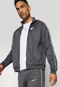 Grå atletisk jakke med hvide sidelister, prydet med et okse-logo på venstre bryst og et Nike swoosh-logo på højre.
