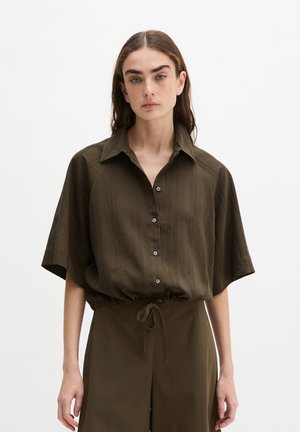 Jeune femme portant une chemise ample à manches courtes olive boutonnée et un pantalon assorti à cordon, debout sur un fond uni.