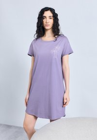 Γυναίκα με σγουρά σκούρα μαλλιά που φοράει ένα άνετο ιώδες φόρεμα T-shirt με κέντημα φοίνικα, στέκεται δίπλα σε μια φωτεινή καρέκλα με υφή.