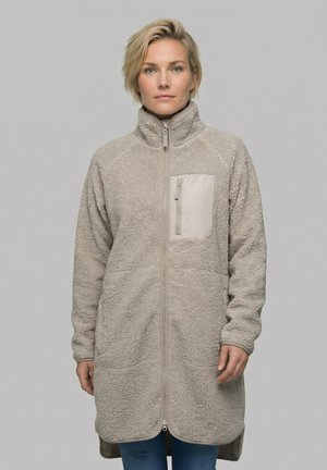 Kvinde iført en lang beige fleecejakke med lynlåsbrystlomme og fuld lynlås foran, stående mod en ensartet lysegrå baggrund.