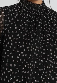 Schwarze Bluse mit hohem Kragen, ausgestattet mit einem verstreuten Blumenmuster in Weiß und Rosa. Der Stoff wirkt leicht und strukturiert.