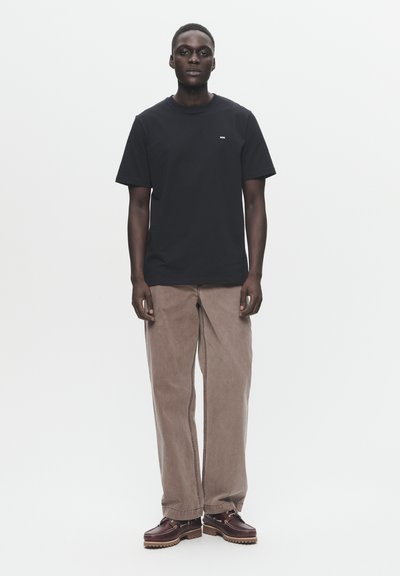 T-shirt preta de algodão com decote redondo e pequeno logotipo branco, combinada com calças largas castanhas claras e sapatos escuros de atar.