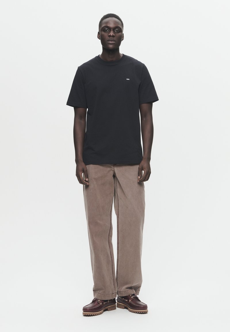 Schwarzes Baumwoll-T-Shirt mit Rundhalsausschnitt und kleinem weißen Logo, kombiniert mit hellbraunen, lockeren Hosen und dunkelbraunen Schnürschuhen.