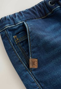 Tumed sinised teksashortsid, millel on tikitud tasku ja pruun nahklabel, millel on välja pressitud tähtede ornament.