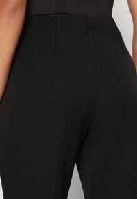 Pantalons noirs sur mesure avec une texture lisse, présentant un détail de couture au dos et une coupe structurée. Le tissu semble léger et polyvalent.