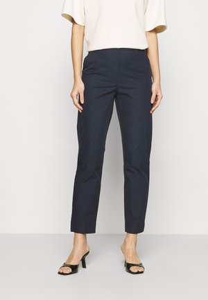 Pantaloni blu navy con vestibilità su misura, tessuto morbido, lunghezza cropped sopra la caviglia, abbinati a tacchi aperti neri.