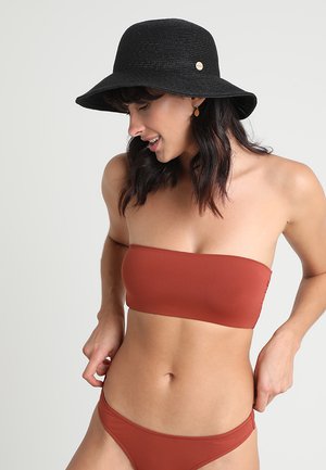 Femeie într-un bikini fără bretele de culoare ruginiu, ajustând partea de jos, purtând o pălărie neagră cu boruri largi și cercei auriu candelabru, uitându-se în jos și zâmbind.