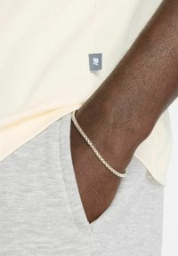T-shirt en coton jaune clair avec une petite étiquette logo grise. Shorts en coton gris et un bracelet en métal argenté sur un poignet à la peau brune.