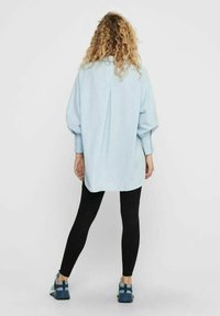 Camicia oversize azzurro chiaro con maniche a sbuffo, dettaglio di piega sulla schiena, abbinata a leggings neri e scarpe da ginnastica blu.