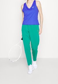 Top deportivo sin mangas púrpura con escote en V, combinado con pantalones verdes ajustados. Sosteniendo una raqueta de tenis blanca. Zapatillas deportivas blancas.