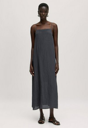 Graues, knöchellanges Kleid mit dünnen Trägern und einem strukturierten, zerknitterten Stoff. Getragen mit schwarzen Riemchensandalen, stehend vor einem neutralen Hintergrund.