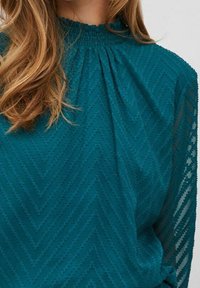 Blusa turchese con un motivo zigzag testurizzato, scollatura arricciata e maniche lunghe trasparenti. Il tessuto ha una leggera lucentezza e una morbida caduta.