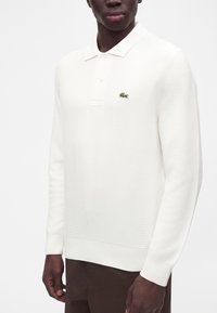 Homme portant un polo blanc à manches longues avec un petit logo crocodile vert sur la poitrine gauche et un pantalon foncé.